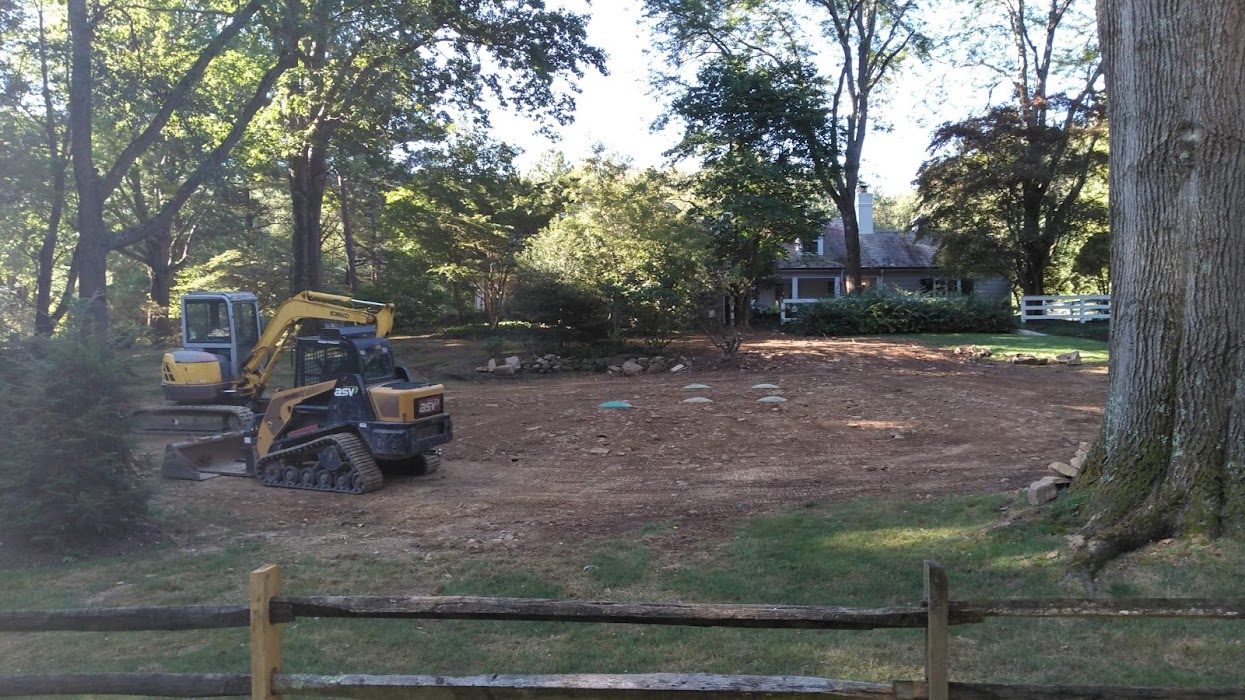 A loader truck and excavator vehicle parked to the side of an old house with a wide woodland property